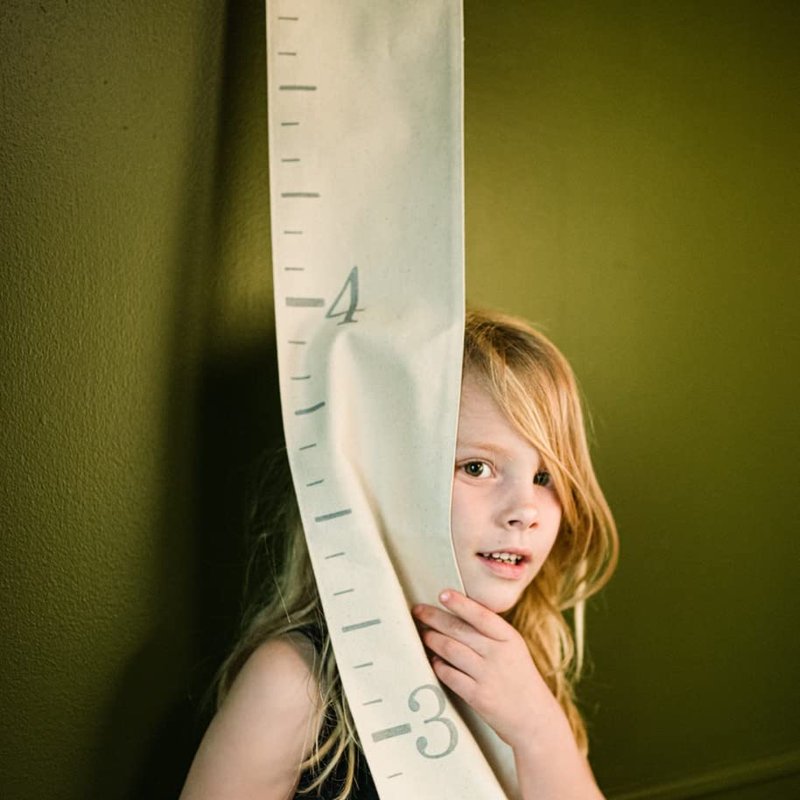 A child peeks out from behind a cream canvas growth chart with gray ruler numbers hanging on a wall