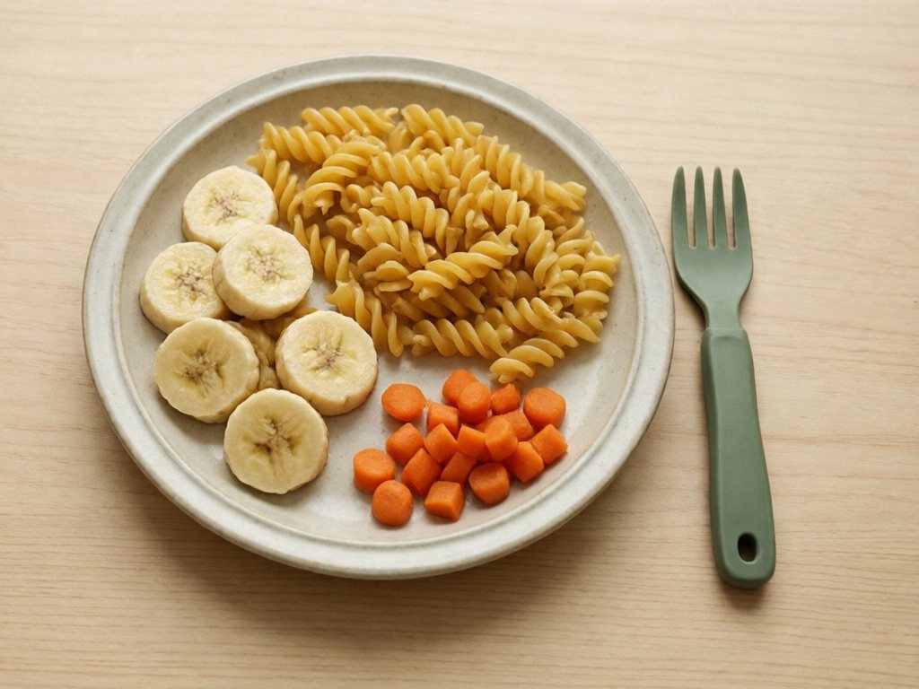 Plate of banana coins, rotini, and soft carrot pieces with a short blunt silicone toddler fork